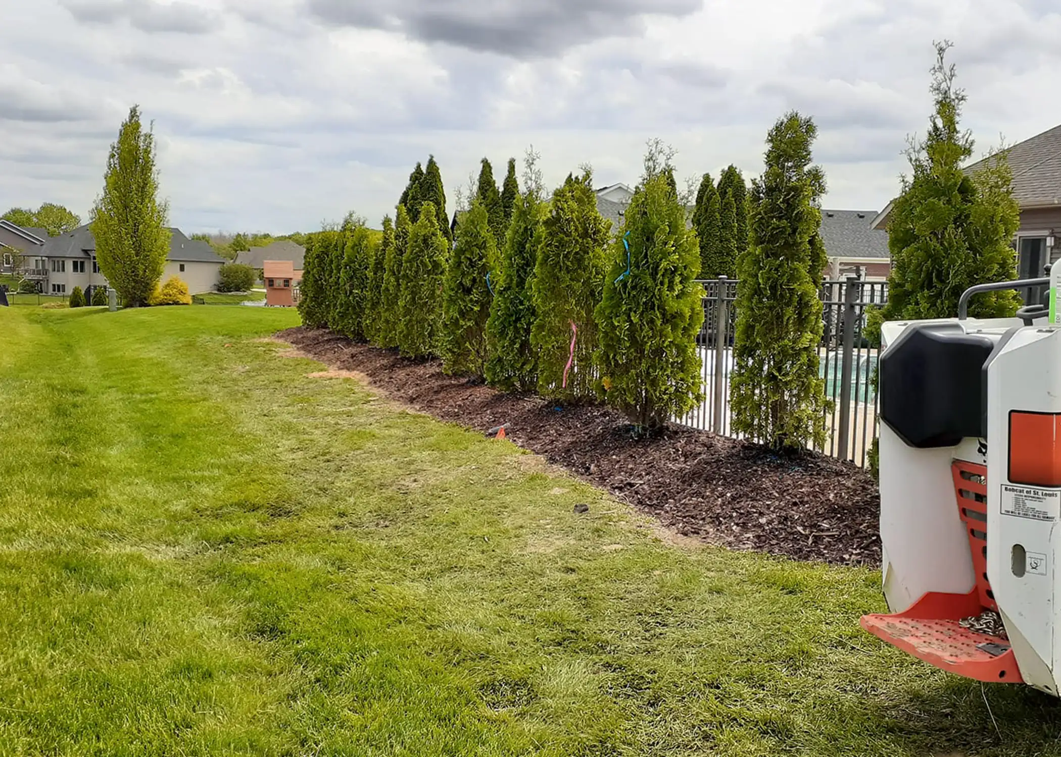 A freshly installed row of Arborvitae view block trees lines a pool fence in a suburban backyard, with a Bobcat skid steer nearby after completing the landscaping work, delivered and planted by Paul Tree Farms in Pleasant Plains, IL